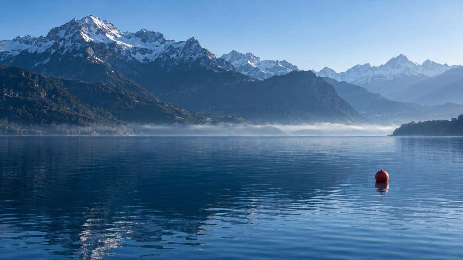 Ruhiger Schweizer Bergsee im Morgenlicht, tiefblaues Wasser und blaue Bergschatten, ein Nebelband vor dem bewaldeten Ufer, eine einzelne rote Boje als einziger warmer Farbakzent
