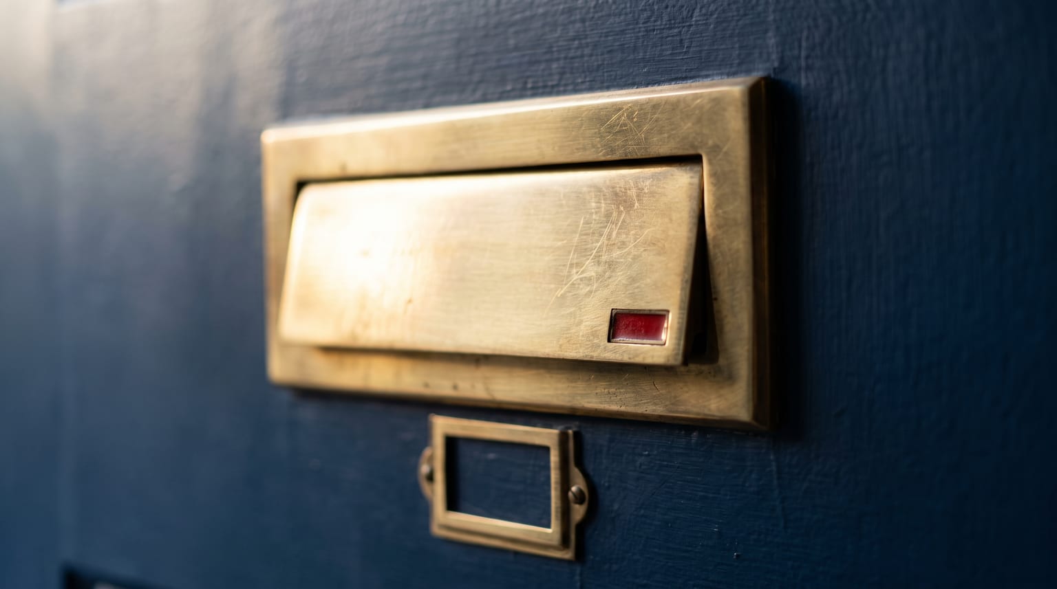 Nahaufnahme einer geschlossenen Messing-Briefkastenklappe an einer dunkelblauen Wand, mit einem kleinen roten Detail
