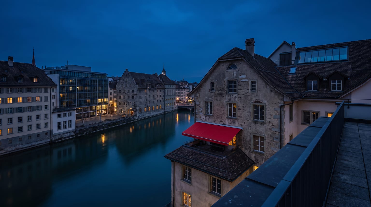 Blick über eine Schweizer Altstadt am Fluss zur blauen Stunde, Spiegelungen im ruhigen Wasser, warme Fensterlichter, eine rote Markise als einziger starker Farbakzent