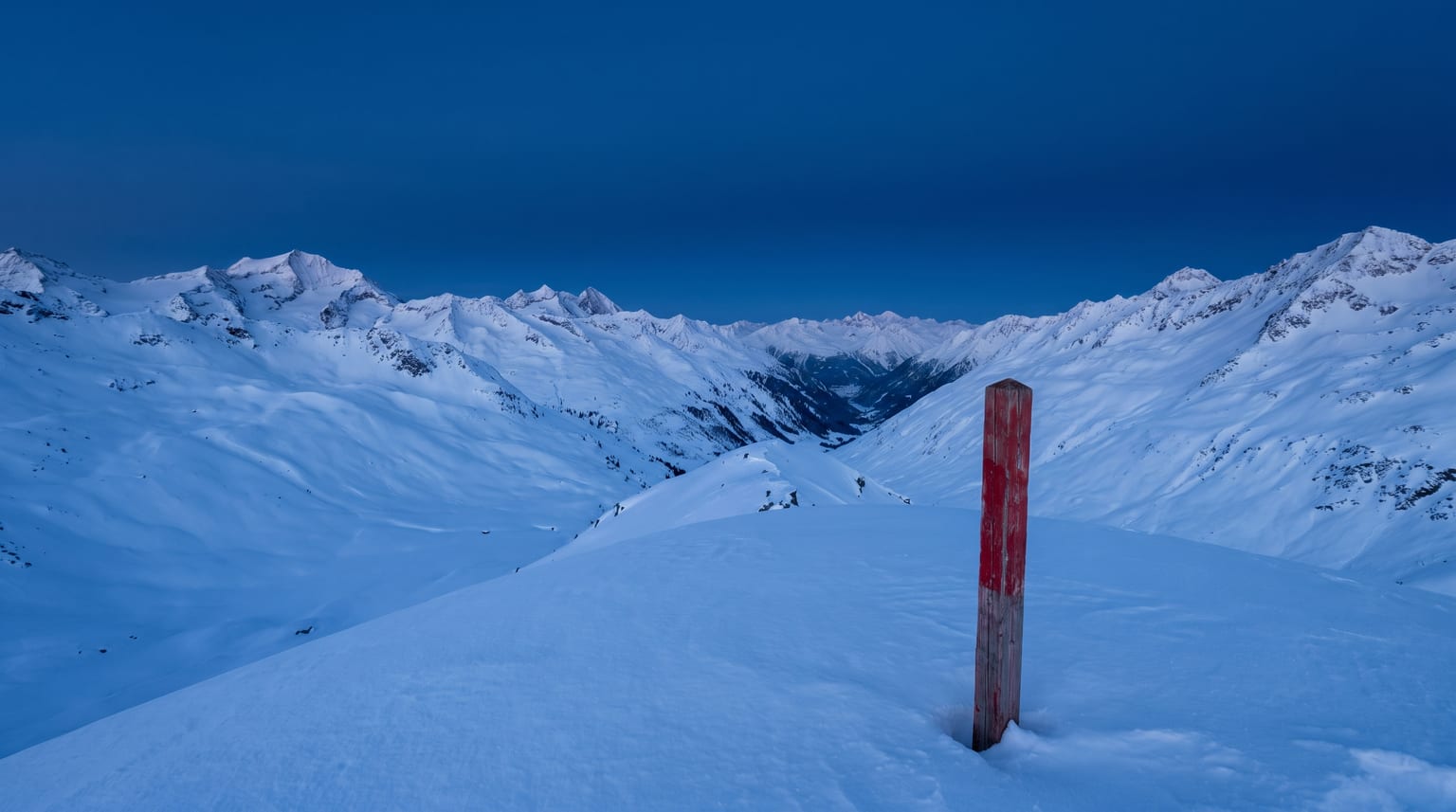 Verschneites Schweizer Bergtal im blauen Abendlicht, ein verwitterter roter Markierungspfahl im Vordergrund